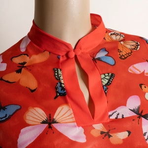 May include: A red blouse with a mandarin collar and a keyhole neckline. The blouse is covered in a pattern of colorful butterflies.
