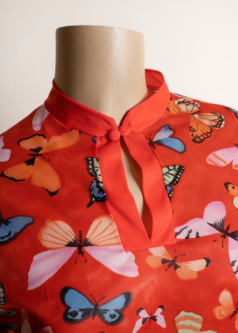 May include: A red blouse with a mandarin collar and a keyhole neckline. The blouse is covered in a pattern of colorful butterflies.