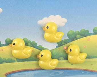 May include: Four yellow duck figurines with orange beaks and black eyes are arranged on a painted background of a pond, grass, and sky. One duck is near a white cloud. The ducks are a bright yellow color.