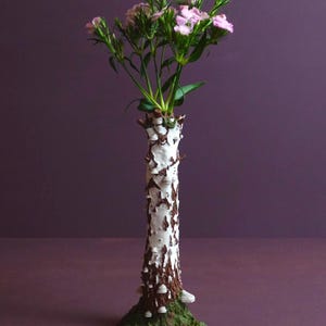Könnte beinhalten: Eine hohe, zylindrische Vase, die wie ein weißer Birkenstamm gestaltet ist, mit strukturierten braunen Rindenflecken und kleinen weißen Baumpilzen. Der Sockel ist mit einer grünen moosartigen Textur bedeckt und enthält rosa Blumen.