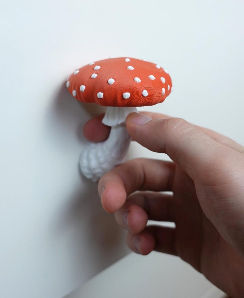 May include: A red and white mushroom with white polka dots. The mushroom has a white stem and is held by a hand. The mushroom is a decorative item.