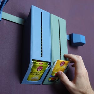 May include: Two blue and green wall-mounted tea bag dispensers with a slot for dispensing tea bags. The dispensers are filled with tea bags labeled "Fenchel-Anis Kümmel" and "Earl Grey".