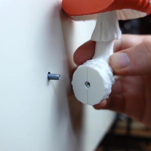 May include: A decorative mushroom-shaped wall hook with a red cap and white spots. The hook is white and is being installed with a screw into a white wall. The mushroom is held by a person's hand.