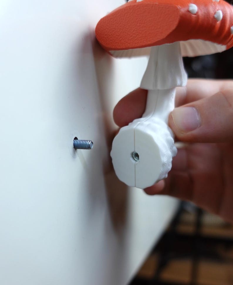 May include: A decorative mushroom-shaped wall hook with a red cap and white spots. The hook is white and is being installed with a screw into a white wall. The mushroom is held by a person's hand.