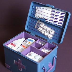 May include: A blue plastic first aid box with a purple handle and cross-shaped accents. The open lid features purple elastic bands holding medical supplies. The interior contains multiple compartments filled with various medicine bottles and blister packs.