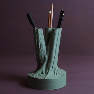 Puede incluir: Un organizador de escritorio de cerámica verde con forma de tronco de árbol con tres ranuras para bolígrafos y lápices. El organizador tiene una superficie texturizada y una base redonda.