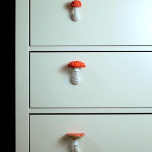 May include: A close-up of a white dresser with three drawers, each adorned with a mushroom-shaped knob. The knobs feature a red cap with white spots and a white stem. The dresser is painted in a light cream color.