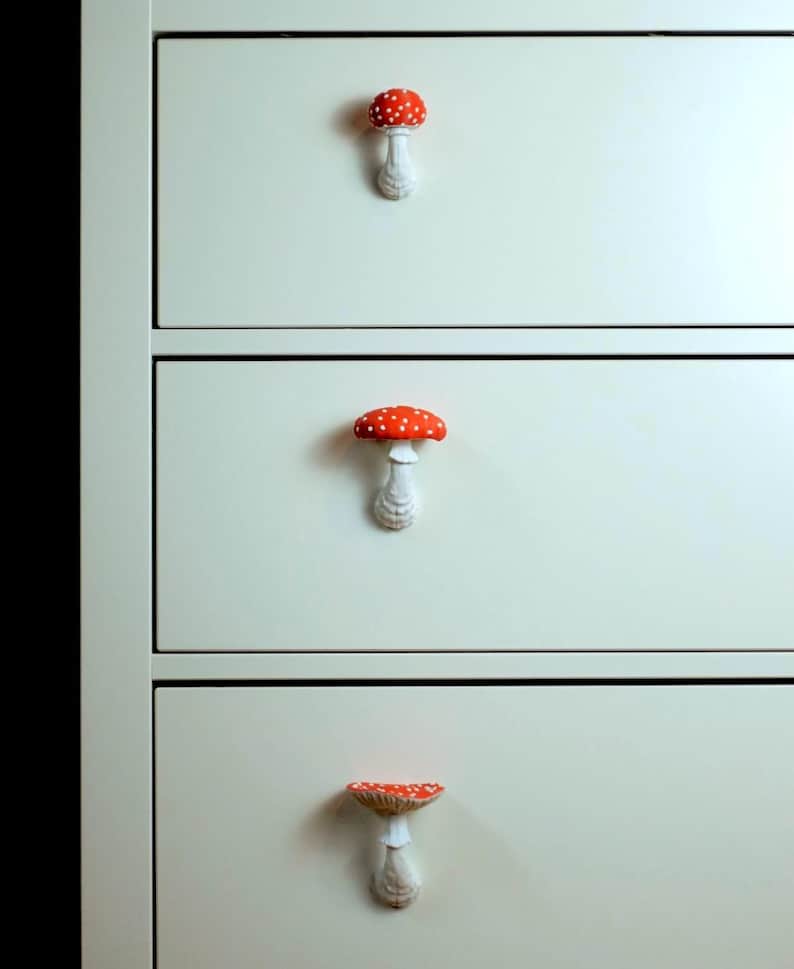 May include: A close-up of a white dresser with three drawers, each adorned with a mushroom-shaped knob. The knobs feature a red cap with white spots and a white stem. The dresser is painted in a light cream color.
