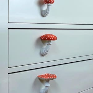 May include: White dresser with three drawers, each adorned with a whimsical mushroom-shaped knob. The knobs feature a red cap with white spots and a white stem, adding a touch of fantasy to the furniture.