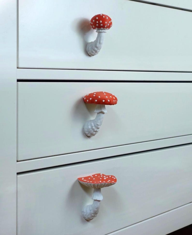May include: White dresser with three drawers, each adorned with a whimsical mushroom-shaped knob. The knobs feature a red cap with white spots and a white stem, adding a touch of fantasy to the furniture.