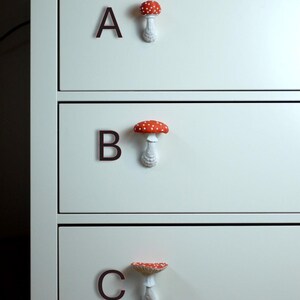 May include: A close-up of a white dresser with three drawers. Each drawer has a dark brown letter and a mushroom-shaped knob. The mushroom knobs have red caps with white spots and white stems. The letters are A, B, and C.