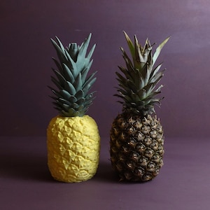 Könnte beinhalten: Zwei Ananas, eine mit einer gelben lackierten Außenseite und die andere mit einer natürlichen braunen Außenseite, werden vor einem violetten Hintergrund gezeigt.