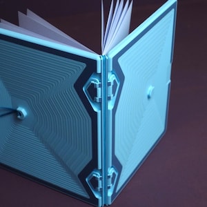 Może przedstawiać: Jasnoniebieski notatnik ze sztywną, teksturowaną okładką z geometrycznym wzorem koncentrycznych linii. Okładka składa się z dwóch zawiasowych paneli połączonych centralnym grzbietem, z widocznymi wewnątrz białymi kartkami papieru.
