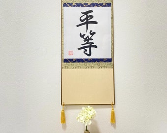 Puede incluir: Un rollo japonés tradicional con la palabra "Año" escrita en caligrafía negra sobre un fondo blanco. El rollo está montado en una tela beige con borlas doradas y un borde decorativo azul y dorado. El rollo está colgado en una pared blanca.