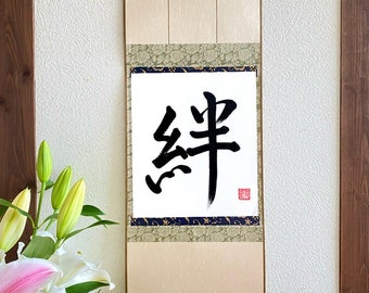 Puede incluir: Un rollo japonés tradicional con un carácter de caligrafía negro sobre papel blanco. El rollo está montado sobre un fondo beige con un borde decorativo y borlas doradas.