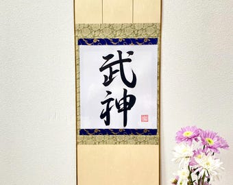 Puede incluir: Un rollo japonés tradicional con los caracteres "武神" (Bushin) escritos en tinta negra sobre papel blanco. El rollo está enmarcado en madera con borlas doradas y está montado en una pared beige. Un ramo de flores rosas y blancas está en un jarrón de vidrio transparente sobre una mesa de madera.
