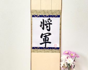 Puede incluir: Un rollo japonés tradicional con marco de madera y borlas doradas. El rollo presenta una inscripción de caligrafía negra sobre papel blanco con los caracteres "静寂" (seijaku) que significa "tranquilidad" o "silencio".