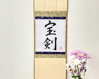 Puede incluir: Un rollo japonés tradicional con los caracteres "金剛" escritos en tinta negra sobre papel blanco. El rollo está montado en un marco de madera con borlas doradas. El rollo está colgado en una pared blanca con un jarrón de flores rosas y blancas delante.