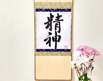 Puede incluir: Un rollo japonés tradicional con los caracteres "青龍" (Seiryu) escritos en tinta negra sobre papel blanco. El rollo está montado en un marco de madera con borlas doradas. El rollo está colgado en una pared blanca con un jarrón de flores rosas y blancas delante.
