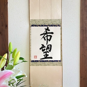 Puede incluir: Un rollo japonés tradicional con la palabra "Esperanza" escrita en caligrafía negra. El rollo tiene un fondo color crema con bordes decorativos y borlas doradas. Un ramo de lirios y otras flores está en primer plano.