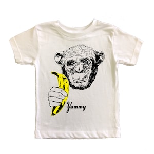Puede incluir: Camiseta blanca con una ilustración en blanco y negro de un mono sosteniendo un plátano amarillo. El texto "Yummy" está impreso debajo del mono.