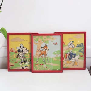 May include: Three framed vintage Disney prints featuring Mickey Mouse, Bambi, and Donald Duck. Each print has a red frame and depicts the characters in colorful scenes. The prints are displayed on a white surface.