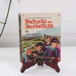 Może przedstawiać: Vintage "Bedknobs and Broomsticks" Little Golden Book. Okładka przedstawia ilustracje dzieci na pojeździe, z tytułem w kolorze czerwonym i pomarańczowym. Książka jest prezentowana na ciemnobrązowym drewnianym stojaku.