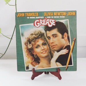 Può includere: Un disco in vinile vintage della colonna sonora del film "Grease" con John Travolta e Olivia Newton-John. La copertina dell'album è verde con il titolo del film in rosso e i nomi degli attori in giallo. Una matita è appoggiata sull'album.