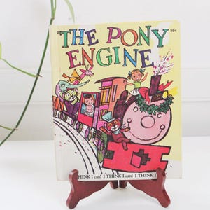 May include: A vintage children's book titled "The Pony Engine" is displayed on a small wooden easel. The book cover features a colorful illustration of a train with passengers and the text "THE PONY ENGINE". The easel has the text "HINK I can! I THINK I can! I THINK I".