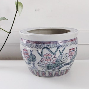 May include: A round ceramic planter with a white base, adorned with a floral design in pink, blue, and green. The planter has a wide rim and a decorative border. A green plant with leaves is visible in the background.