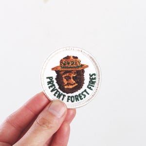 Może przedstawiać: Okrągła, haftowana naszywka przedstawiająca Smokey Bear w kapeluszu strażnika. Naszywka ma napis "PREVENT FOREST FIRES" w kolorze zielonym na obrzeżu. Niedźwiedź jest brązowy, a kapelusz brązowy. Naszywka jest biała z wyszywaną obwódką.