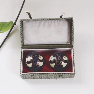 Puede incluir: Un par de bolas decorativas azul oscuro con diseños de panda dentro de una caja verde estampada. La caja tiene un interior rojo y un forro de tela blanca. Las bolas son redondas y presentan ilustraciones de pandas en blanco y negro.