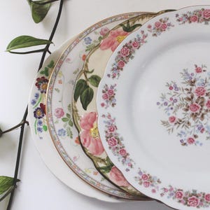 Może przedstawiać: Stos trzech dekoracyjnych talerzy porcelanowych z kwiatowymi wzorami. Talerze zdobią różowe róże, inne kwiaty i złote obramowania. Zielona winorośl z liśćmi znajduje się na pierwszym planie.