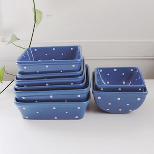 Puede incluir: Un conjunto de cuencos cuadrados de cerámica azul con lunares blancos. Los cuencos, de varios tamaños, están apilados, con un juego a juego a la derecha. Se muestran sobre una superficie blanca.