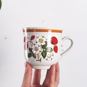 Può includere: Una tazza in ceramica bianca con bordo e manico marroni, decorata con un motivo a fragole e fiori. La tazza è tenuta in mano su sfondo bianco. Le fragole sono rosse e i fiori bianchi con centri gialli.