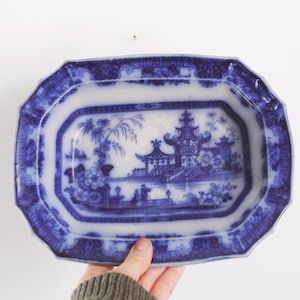 Puede incluir: Una fuente de porcelana azul y blanca antigua. La fuente rectangular presenta una escena de paisaje detallada con una pagoda y elementos florales. El borde tiene un borde decorativo. Una mano sostiene la fuente.