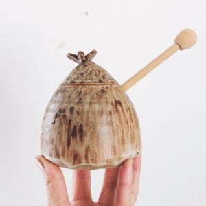 May include: A handmade ceramic honey pot with a wooden honey dipper. The pot is a light brown color with darker brown streaks and a decorative floral-shaped lid. The dipper is light-colored wood.