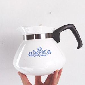 Könnte beinhalten: Weiße Keramik-Kaffeekanne mit schwarzem Griff und silbernem Band am oberen Rand. Die Kanne hat ein blaues Blumendesign. Die Kaffeekanne wird vor einem weißen Hintergrund gehalten.