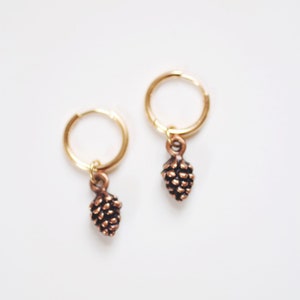 May include: A pair of gold hoop earrings with small, detailed pine cone charms hanging from them.