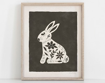 May include: A black and white illustration of a rabbit with floral details on a dark brown background. The rabbit is sitting with its ears up and its eyes closed. The illustration is framed in a light wood frame.