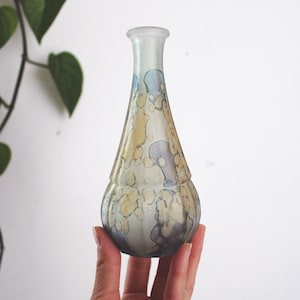 May include: A decorative glass vase with a bulbous base and a slender neck. The vase features a pattern of muted blue, yellow, and gray splotches. The vase is held in a hand against a white background.