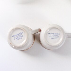 May include: Two off-white stoneware mugs with handles, featuring the text "strawberries n cream" and "STONEWARE JAPAN" in blue. The mugs are set against a white background with a green plant.
