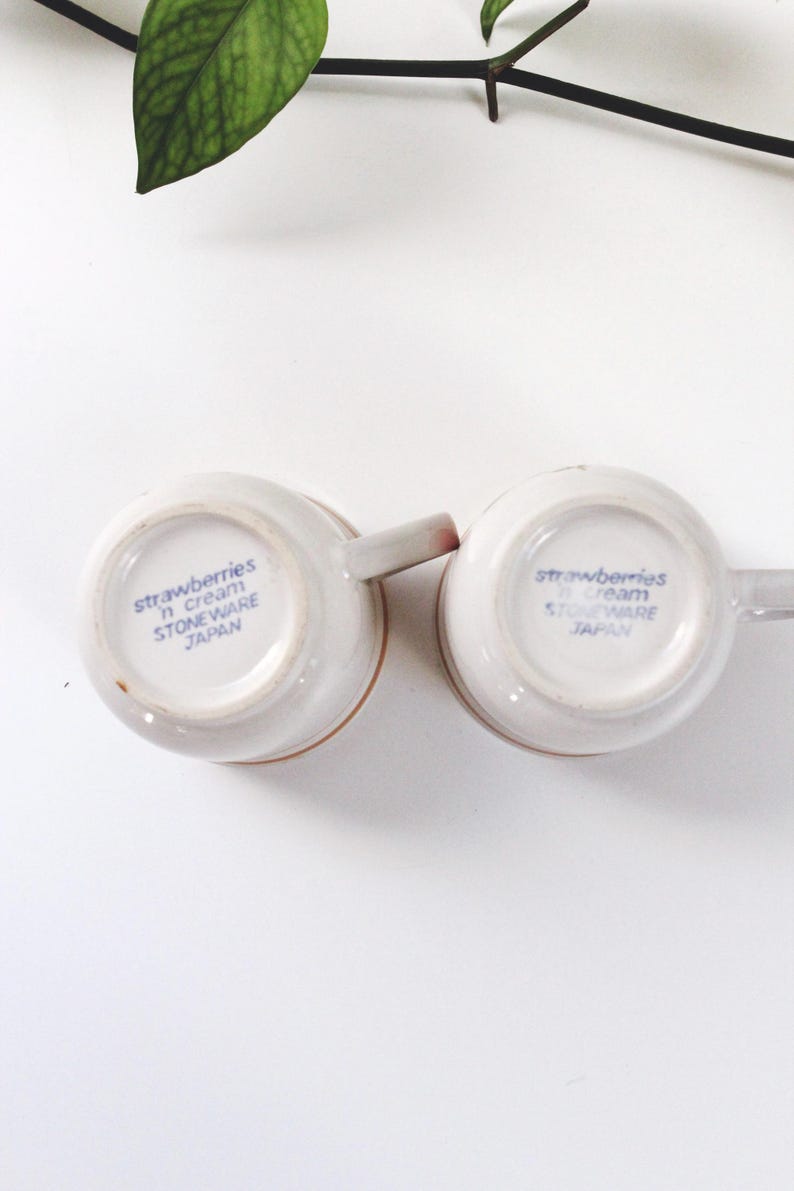 May include: Two off-white stoneware mugs with handles, featuring the text "strawberries n cream" and "STONEWARE JAPAN" in blue. The mugs are set against a white background with a green plant.