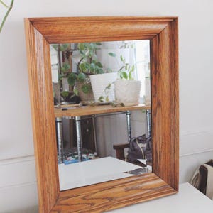May include: A rectangular wall mirror featuring a thick, medium-toned brown wood frame with a visible natural grain pattern. The frame has a beveled edge design that slopes inward toward the reflective glass surface, which shows a reflection.
