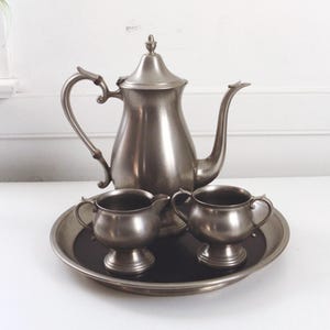Puede incluir: Juego de té de peltre vintage, que incluye una tetera, una jarra de crema, un azucarero y una bandeja. El juego tiene un diseño clásico con asas curvas y un acabado pulido. La bandeja es redonda.