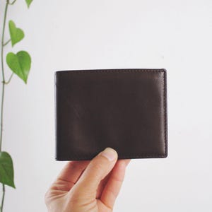 May include: A dark brown leather bifold wallet with a smooth, matte finish and clean, straight stitched edges. The wallet is rectangular in shape and features a simple, minimalist design with no visible branding or external hardware.