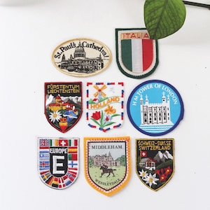 May include: A collection of embroidered patches featuring various travel destinations. Patches include St. Paul's Cathedral, the Tower of London, and flags representing Europe, Italy, and Switzerland. Other patches depict Holland, Liechtenstein, and Middleham.