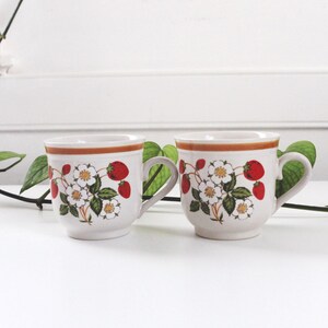May include: Two white ceramic mugs with a strawberry and floral design. Each mug features a brown rim and handle, with red strawberries, white flowers, and green leaves. The mugs are set against a white background.