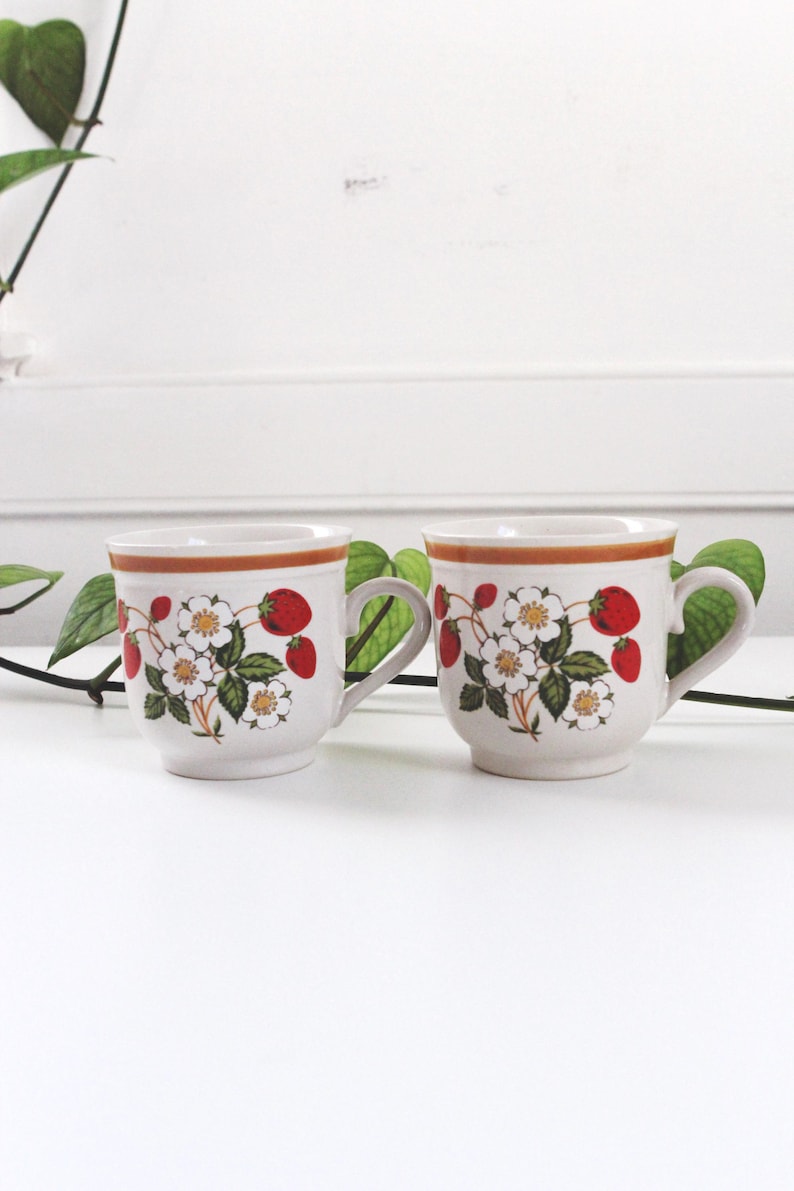 May include: Two white ceramic mugs with a strawberry and floral design. Each mug features a brown rim and handle, with red strawberries, white flowers, and green leaves. The mugs are set against a white background.