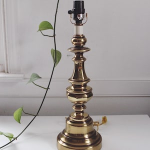 Puede incluir: Una lámpara de mesa de latón vintage con un diseño clásico y ornamentado. La lámpara tiene un portalámparas en la parte superior y una base redonda. Una vid verde con hojas está en el fondo. La lámpara está sobre una superficie blanca.